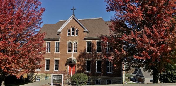 A large brick church building with arched windows and a cross on the roof stands amid vibrant red-leaved trees under a clear blue sky. A sign is visible near the entrance.