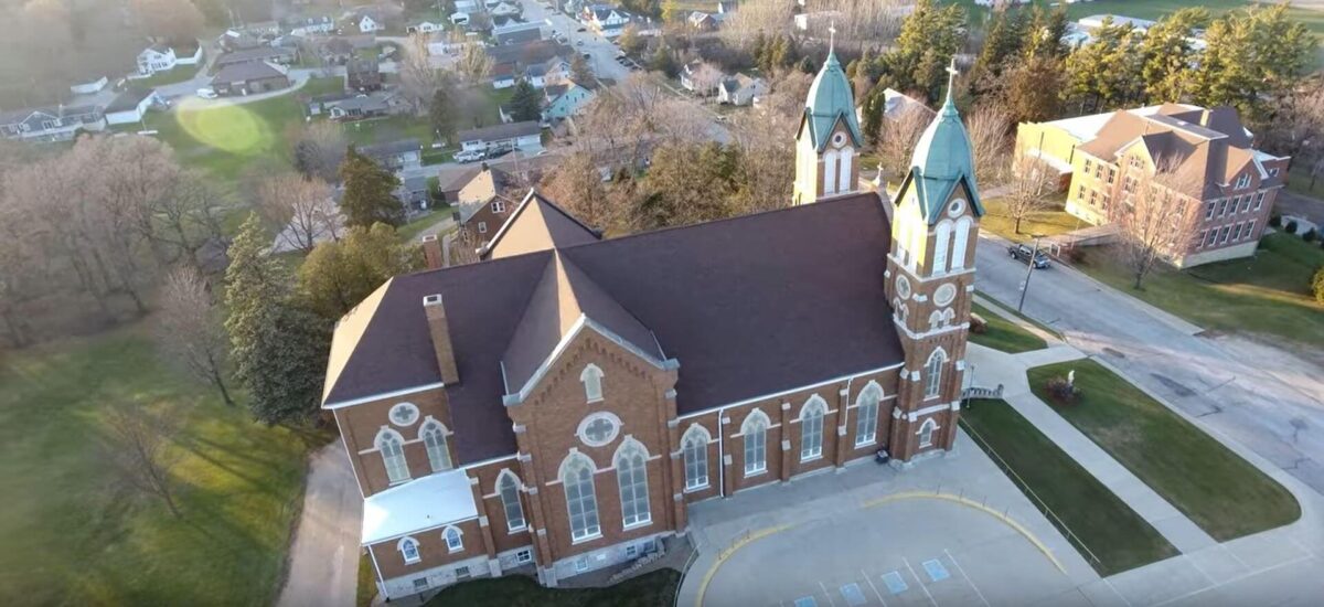 Aerial view of a large brick church with green-roofed towers, surrounded by trees and a residential neighborhood. Paved roads and sidewalks are visible, along with another brick building nearby.