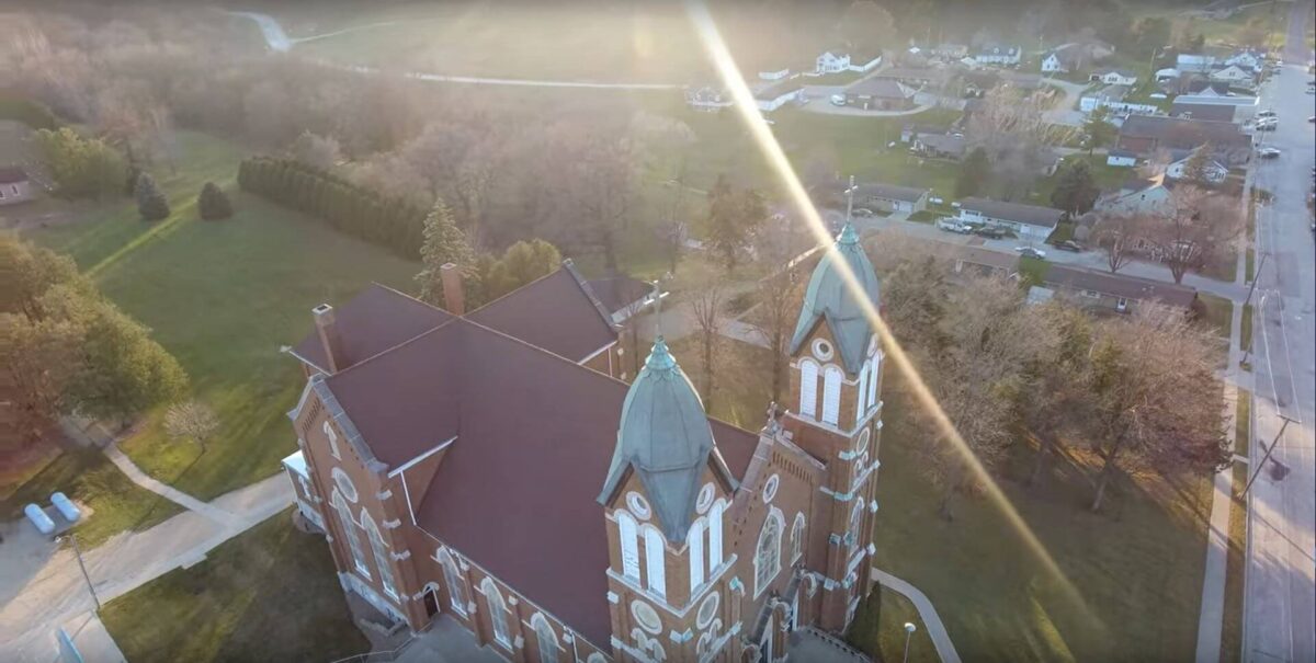 Aerial view of a large brick church with two towers, surrounded by trees and houses. Sunlight creates lens flares across the image, with a road visible to the right and open green spaces around the church.