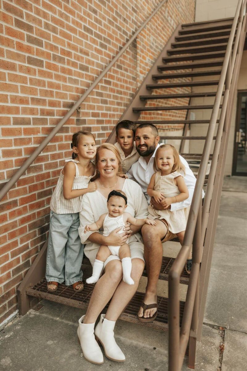 A family of six sits on outdoor stairs. A woman holds a baby, while a man sits beside them. Three children stand and sit near the adults, smiling. They're against a brick wall. The atmosphere is warm and casual.